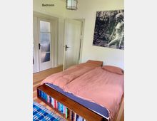 Schlafzimmer mit einem Doppelbett mit altrosa Bettwäsche, darunter ein integriertes Bücherregal mit verschiedenen Büchern. Im Hintergrund sind eine halb geöffnete Tür und eine Glastür sowie ein großes Schwarz-Weiß-Bild an der Wand zu sehen.