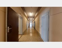 Langer, schlichter Flur in einem Gebäude mit beigefarbenen Wänden und hellbraunem Laminatboden, gesäumt von weißen Türen, von denen einige geöffnet sind, während Neonröhren die Decke beleuchten. Am Ende des Flurs ist ein heller Lichtschein sichtbar.