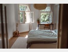 Helles Schlafzimmer mit weiß gestrichenen Wänden, einem Bett mit hellgrüner Bettdecke, einem Holztisch und Stuhl vor großen Fenstern, die Blick auf Bäume bieten. Eine kleine Kiste mit einer Vase steht neben dem Bett, und ein runder Papierlampenschirm hängt von der Decke.