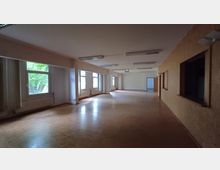 Großer, leerer Raum mit Holzfußboden, weißen Wänden und mehreren Fenstern, die einen Blick auf grüne Bäume bieten. An der rechten Wand befinden sich mehrere rechteckige Öffnungen, links sind Heizkörper unter den Fenstern installiert, und an der Decke sind rechteckige Leuchten angebracht.