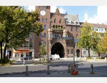 Backsteingebäude mit historischem Torbogen und Turm mit Uhr, umgeben von Bäumen und Straßenlaternen in einem städtischen Umfeld; Straßenschilder weisen auf die Kreuzung von Schönhauser Allee und Wörther Straße hin.