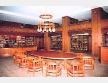 Ein holzvertäfelter Bibliotheksraum mit hohen Bücherregalen entlang der Wände, einem ovalen Tisch in der Mitte, umgeben von Holzstühlen, und einem großen, mehrarmigen Kronleuchter an der Decke.