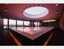 Modernes Spa mit einem beleuchteten Pool, eingerahmt von einer Glasfassade, die eine Aussicht auf eine Landschaft bietet. Die Decke hat eine große runde Öffnung und trägt den Schriftzug „Heaven“; die Beleuchtung erzeugt warme Farbakzente.