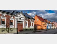 Straßenszene mit einer Reihe von Wohnhäusern in unterschiedlichen Farben und architektonischen Stilen, darunter Fachwerk- und Backsteinelemente. Vor den Gebäuden stehen junge Bäume, und auf manchen Dächern sind Solarpaneele sichtbar; der Himmel ist bewölkt.