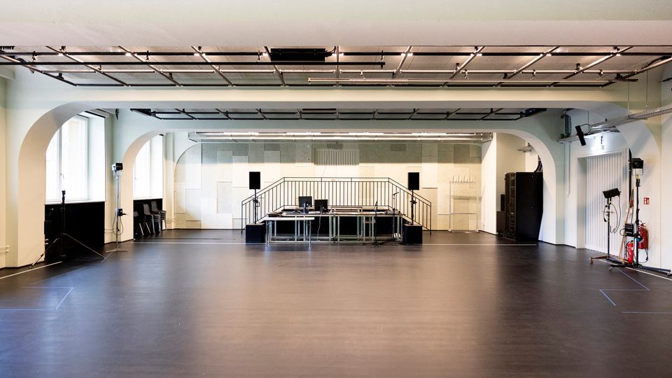Großer, leerer Studio- oder Proberaum mit dunklem Boden, weiß gestrichenen Wänden und breiten Rundbögen. Im Hintergrund steht eine kleine Bühne mit Tischen, Monitoren und Lautsprechern; an der Decke sind Schienen mit Beleuchtung und Technik montiert, seitlich befinden sich Fenster und Equipment.