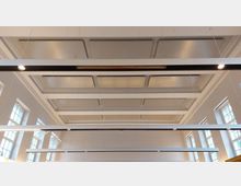 Großer Innenraum mit einer klassischen Kassettendecke und modernen, länglichen Hängeleuchten. Auf beiden Seiten sind hohe Fenster mit Rahmen im rechteckigen Stil angeordnet.