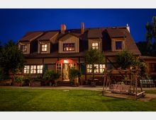 Fachwerkhaus mit Satteldach und mehreren Dachgauben, beleuchtet in der Abenddämmerung. Vor dem Haus befinden sich eine Holzschaukel, Bäume, Pflanzen sowie gestapelte Holzscheite.