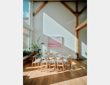 Heller Innenraum mit hohen weißen Wänden und Holzträgern, eingerichtet mit einem Glastisch, umgeben von Holzdrehstühlen mit gepolsterten Sitzflächen. Große Fenster lassen viel Tageslicht herein, Pflanzen und ein gerahmtes Bild schmücken den Raum.