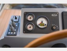 Nahaufnahme eines Steuerpults mit Bedienelementen eines Schiffs. Sichtbar sind ein Zündschlüssel, Drehzahlmesser, Druckanzeigen sowie zwei Steuerhebel; der Hintergrund zeigt verschwommen einen Holzboden und Wasser.