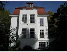 Weiße Villa mit rotem Walmdach und mehreren hohen Fenstern mit schlichtem Metallgeländer, umgeben von Bäumen und Schatten. Das Gebäude steht in einer grünen Gartenumgebung bei klarem Himmel.