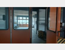 Innenansicht einer verlassenen Industrieanlage mit orangefarbenen Schiebetüren. Im Hintergrund sind große Fenster, Wellblechwände und abgenutzte Böden zu sehen, mit einem gelb-schwarz gestreiften Betonsockel im Vordergrund.