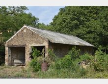 Backsteingebäude mit abblätterndem Wellblechdach, umgeben von wilder Vegetation und einigen Bruchsteinen, am Rand eines bewaldeten Bereichs gelegen.