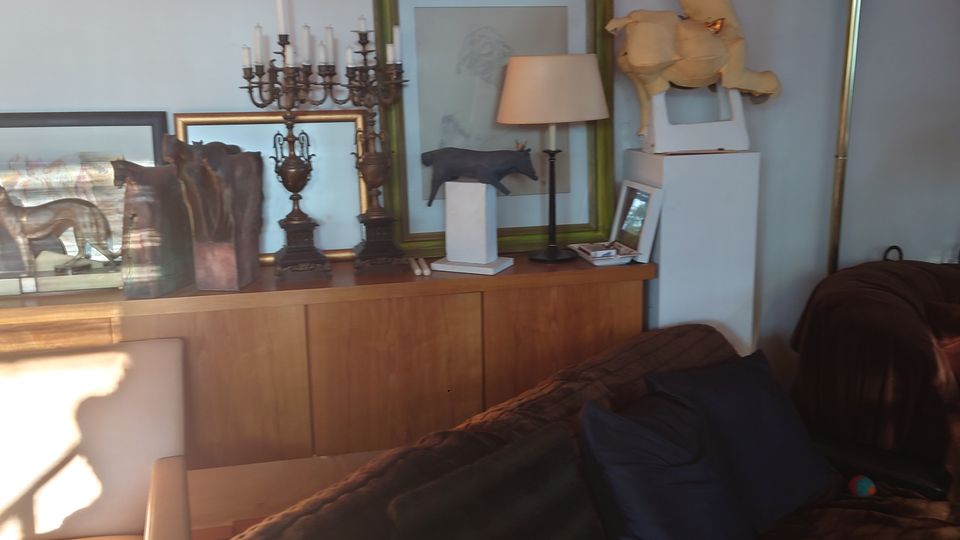 Wohnzimmer mit einer braunen Couch, Kissen und einer Holzkommode an der Wand. Auf der Kommode stehen dekorative Gegenstände wie ein mehrarmiger Kerzenständer, Bilderrahmen, eine Tischlampe und ein kleines Modell eines Pferdes.