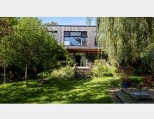 Rückansicht eines modernen Hauses mit Holzfassade und großen Glasfenstern, umgeben von einem üppig bewachsenen Garten mit Rasen, Sträuchern und Bäumen. Eine Terrasse mit Treppe führt von der leicht erhöhten Gebäudeeingangsebene in den Garten.