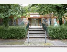 Backstein-Gebäude mit dem Schild „Freizeitstätte Koeltzepark“ über dem Eingang, umgeben von Bäumen und Sträuchern. Die Wände sind mit Graffiti bedeckt, und ein gepflasterter Weg führt über mehrere Stufen zum Eingang.