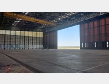 Große, leere Flugzeughalle mit einer hohen Decke aus Stahlträgern und einem geöffneten Hangartor, das den Blick ins Freie mit blauem Himmel und Bäumen am Horizont freigibt. Im Vordergrund sind Feuerlöscher und einige Personen in Schutzkleidung erkennbar.