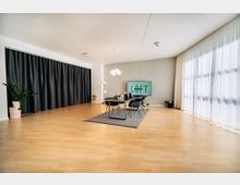 Moderner, heller Besprechungsraum mit Holzfußboden, einem zentralen Tisch mit schwarzen Stühlen auf einem grauen Teppich und einem großen Bildschirm an der Wand mit der Aufschrift „LÖFT“. Links schwarze Vorhänge und eine Pflanze, rechts große Fenster mit weißen Vorhängen.