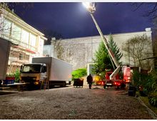 Ein nächtlicher Außenbereich mit einem weißen Transport-Lkw, einer Hebebühne und Arbeitsgeräten. Im Hintergrund sind eine Hausfassade mit Gerüst, eine mit Efeu bewachsene Wand und einige Bäume sichtbar, Beleuchtung sorgt für Helligkeit in der Szene.