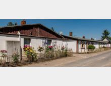 Reihenhäuser mit weiß gestrichenen Fassaden und teils hölzernen Giebeln, davor kleine Vorgärten mit Blumen und einem niedrigen Drahtzaun, im Hintergrund schmaler Gehweg und blauer Himmel.
