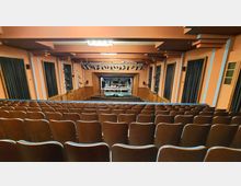 Innenraum eines Theatersaals mit mehreren Sitzreihen aus Holzstühlen, die zur Bühne führen. Die Bühne ist von holzvertäfelten Wänden und schwarzen Vorhängen eingefasst, während an der Decke Bühnenscheinwerfer montiert sind.