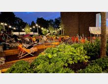 Außenbereich eines Open-Air-Kinos bei Dämmerung, mit Liegestühlen auf Holzplattformen, umgeben von Pflanzenbeeten und Lichterketten. Im Hintergrund ist eine Leinwand an einer dunklen Backsteinwand zu sehen.