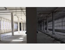 Ein leerer, zweigeteilter Fabrikraum mit Betonpfeilern und einer Rasterdecke. Die Wand auf der linken Seite hat eine Reihe von schmalen Fenstern, während die rechte Seite von großen Glasbausteinfenstern dominiert wird; der Boden ist teilweise beschädigt.