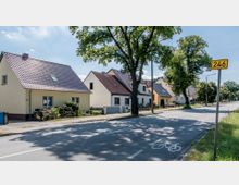 Straßenszene in einem Wohngebiet mit freistehenden Einfamilienhäusern, hellen Fassaden und dunklen Ziegeldächern; am Straßenrand stehen Bäume und ein gelbes Verkehrsschild mit der Nummer 246. Die Straße hat eine markierte Fahrradspur, und der Himmel ist klar bei sonnigem Wetter.