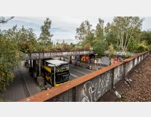 Eine städtische Szene mit einer mobilen Bushaltestelle unter einer rostigen Eisenbahnbrücke, die mit Graffiti bedeckt ist. Umgeben von Bäumen und Plakaten an den Wänden, zeigt das Bild oben Schienen auf der Brücke und einen gelben Doppeldeckerbus der Linie M19 darunter.