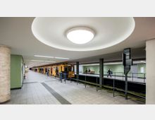 U-Bahn-Station mit modernen Elementen, gelber Zug auf der linken Seite des Bahnsteigs, grüne Wandfliesen im Hintergrund und große runde Deckenleuchte. Menschen laufen und warten auf dem Bahnsteig.