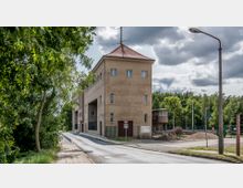 Ein mehrstöckiges Gebäude mit flachem Dach und beige-brauner Fassade, durchbrochen von kleinen Fenstern und großen Durchfahrten, steht an einer schmalen Straße. Im Hintergrund sind Bäume, eine kleine Nebenstruktur mit Panoramafenstern und Bauarbeiten mit Kieshaufen zu sehen.