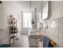 Modernes Badezimmer mit weißen Hochglanzfliesen und grauem Boden. Es gibt ein Waschbecken mit Spiegel, ein Fenster, ein hölzernes Regal mit Handtüchern und Pflegeprodukten sowie eine Wandheizung.