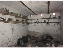 Ein alter Kellerraum mit weiß gestrichenen, rissigen Wänden, einem gewölbten Deckenbogen und kleinen, rechteckigen Fenstern. Holzregale entlang der Wände sind mit verschiedenen Behältern, Flaschen und Gegenständen beladen, während auf dem Boden alte Reifen, Rohre und Werkzeuge verstreut liegen.