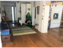 Eingangsbereich eines Wohnhauses mit Holzfußboden, einer offenen Garderobe mit Jacken und Helmen sowie einem gestreiften Teppich. Rechts ist eine moderne Küche mit weißen Schränken und einem eingebauten Backofen zu sehen.