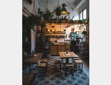 Ein gemütliches Café-Interieur mit Holzmöbeln, hängenden Pflanzen und industriellen Leuchten. Im Hintergrund ist eine Theke mit weiß gefliester Rückwand und Regalen sowie ein Getränkeautomat sichtbar.