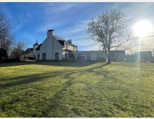 Zweistöckiges, modernes Gebäude mit weißen Wänden und großen Fenstern, umgeben von einer weitläufigen Wiese. Im Hintergrund sind ein kahler Baum, weitere Gebäude und eine kleine Kirche mit Turm zu sehen, während die Sonne tief am Himmel steht.