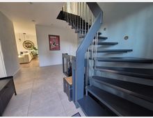 Moderne Inneneinrichtung mit einer offenen Wendeltreppe aus Metall mit dunklen Stufen und silbernen Geländern, auf einem gefliesten Boden. Im Hintergrund ist ein Wohnbereich mit Holzboden, einer hellen Couch, Pflanzen und dekorativen Wandelementen zu sehen.