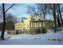 Schlossartige gelbe Villa mit klassizistischer Architektur, bestehend aus einem Hauptgebäude mit zahlreichen Fenstern und einem Säulenpavillon mit Steinfundament. Die Umgebung ist winterlich verschneit, mit kahlen Bäumen und einem zugefrorenen Gewässer im Vordergrund.