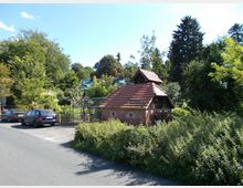 Kleines Ziegelsteingebäude mit Satteldach, umgeben von dichtem Grün und angrenzend an eine schmale Straße mit geparkten Autos; im Hintergrund sind weitere Häuser und große Bäume sichtbar.