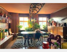 Wohnzimmer mit Vintage-Design und natürlicher Beleuchtung. Ein runder Marmortisch steht auf einem schwarz-weiß gemusterten Teppich, umgeben von farbenfrohen Stühlen und grünen Zimmerpflanzen; an den Wänden befinden sich Regale mit Dekor und ein Sideboard aus Holz.