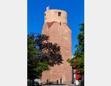 Ein historischer Ziegelturm mit einer runden oberen Etage und kleinen Fenstern, gekrönt von einer Fahne. Der Turm steht in einem städtischen Umfeld, umgeben von Bäumen und modernen Gebäuden.