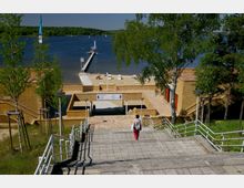 Treppen führen zu einer Uferpromenade mit Sandstrand, einer Anlegestelle und einem Steg, der in einen ruhigen See hinausführt. Umgeben von Bäumen und Gebäuden aus sandfarbenem Mauerwerk, bietet die Umgebung eine Mischung aus Natur und Architektur.