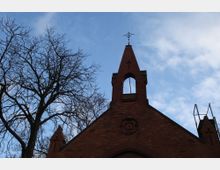 Backstein-Kirchturm mit Spitzdach und geöffnetem Glockenfenster, verziert mit einem Kreuz an der Spitze; im Vordergrund ein laubloser Baum vor blauem Himmel mit Wolken.