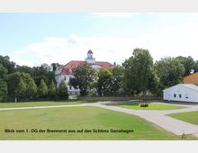 Blick auf das Schloss Genshagen, ein historisches weißes Gebäude mit rotem Walmdach und Turm, umgeben von Bäumen und einer großzügigen Grünfläche. Im Vordergrund sind ein gepflasterter Weg sowie ein modernes, weißes Nebengebäude sichtbar.
