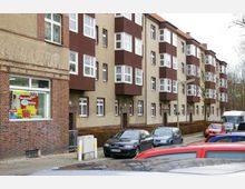 Mehrstöckiges Wohngebäude im Stil der frühen 20. Jahrhunderts mit braunen und beigen Fassaden sowie symmetrischen Erkern. Im Erdgeschoss befindet sich ein Geschäft mit Schaufenster und Werbebanner; verschiedene Fahrzeuge sind entlang der Straße geparkt.