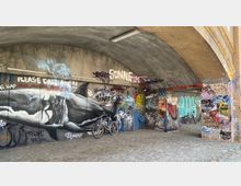 Unterführung mit gemauertem Rundbogen und Wänden, die mit bunten Graffiti und Schriftzügen bedeckt sind, darunter ein großflächiges Haifisch-Motiv. Zwei abgestellte Fahrräder stehen auf dem Kopfsteinpflasterboden.