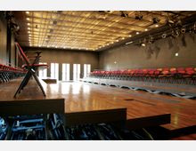 Ein moderner Veranstaltungssaal mit Holzfußboden, ansteigenden Sitzreihen mit roten und orangefarbenen Klappstühlen sowie einer Metall-Decke mit eingebauten Scheinwerfern. Helle Türen auf der Rückseite lassen Tageslicht herein.