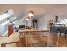 Wohnraum in einem Dachgeschoss mit Holzboden, einer großen Holztafel mit transparenten Stühlen, schrägen Fenstern an der linken Wand und einer offenen Küche im Hintergrund. Moderne Hängelampen und Dekorationselemente wie Pflanzen und Kerzen schmücken den Raum.