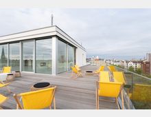 Moderne Dachterrasse mit Holzbelag und Panoramablick auf eine Stadt, ausgestattet mit gelben Liegestühlen, kleinen Rattan-Tischen und einer Glasbrüstung; im Hintergrund ein weißes Gebäude mit großen Fensterfronten.