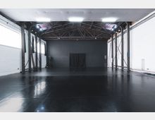 Großer, leerer Raum mit glänzendem schwarzem Boden, weißen Ziegelwänden und offener Holzdecke mit sichtbaren Balken. Im Hintergrund befindet sich eine schwarze Wand mit einem mittig hängenden dunklen Vorhang.