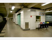 U-Bahn-Station mit Bahnsteig, gebogenem Gleis und moderner Wandverkleidung aus weißen Paneelen; im Vordergrund befinden sich eine Tür, ein Schild mit Verbotszeichen und ein farblich gekennzeichneter Mülleimer für Mülltrennung. Die Decke zeigt freiliegende Kabel und Leuchtstofflampen, und im Hintergrund ist eine Treppe sichtbar.